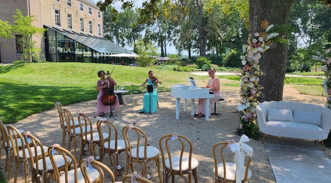 Strijktrio met witte vleugel verzorgt live muziek tijdens een romantische buitenbruiloft met bloemenboog en ceremonieopstelling.
