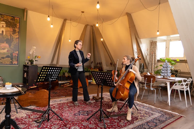 celloleerling speelt cello terwijl de docent uitleg geeft in een lichte leslokaal met lessenaars en een Perzisch tapijt